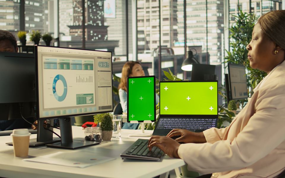 An analyst working across a desktop dashboard, a tablet and a laptop in an open-plan office.